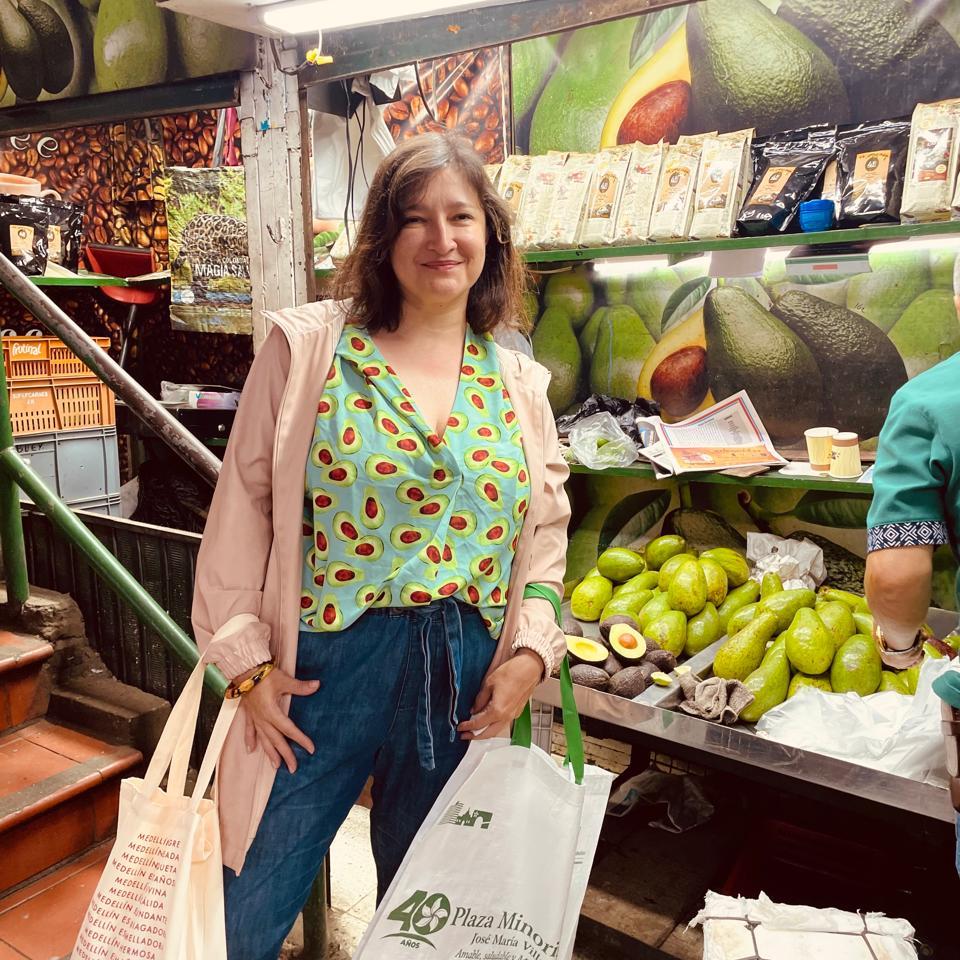 Maya frente a un puesto de aguacates en la Plaza Minorista de Medellín, vistiendo una blusa estampada con aguacates